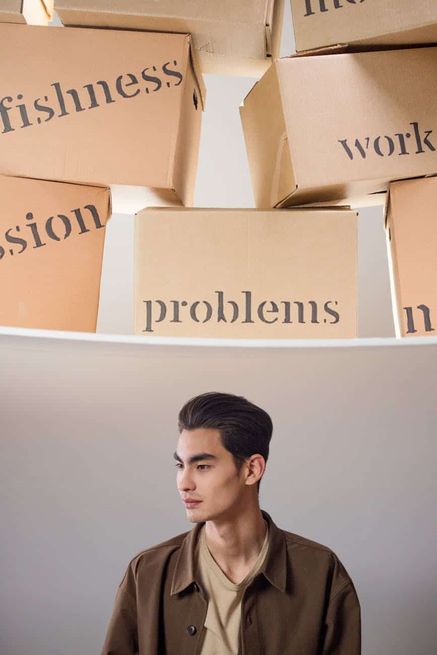 A young man with dark hair and a button-up shirt stands in profile, looking contemplative, with several cardboard boxes labeled with words like 'selfishness', 'problems', 'work', and 'passion' arranged above him.